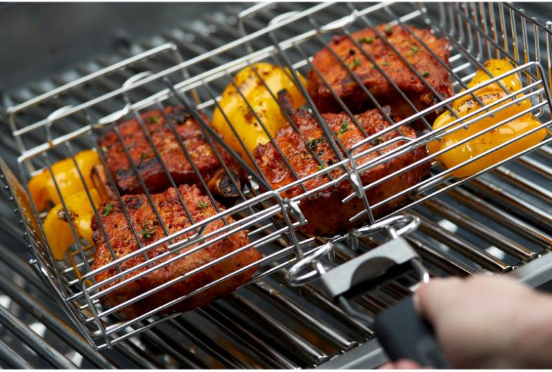 Grilling vegetables and sausages on a metal grill basket with tongs.