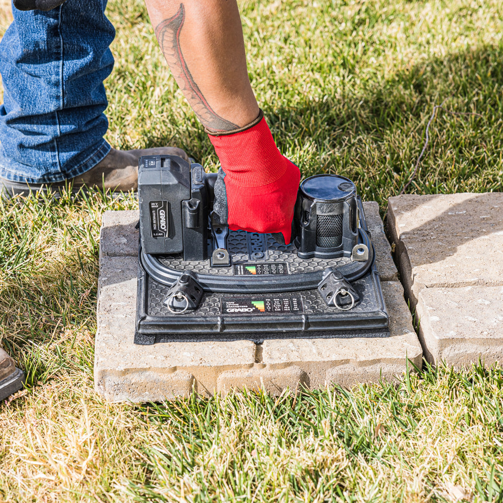 A person wearing blue jeans and a red work glove is operating a black electronic device placed on concrete pavers outdoors on a grassy lawn.