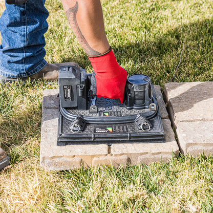 A person wearing blue jeans and a red work glove is operating a black electronic device placed on concrete pavers outdoors on a grassy lawn.