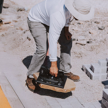 A person in a white long-sleeve shirt, gray pants, and a wide-brim hat operates a black tile leveling machine on a stone slab outdoors on a construction site.