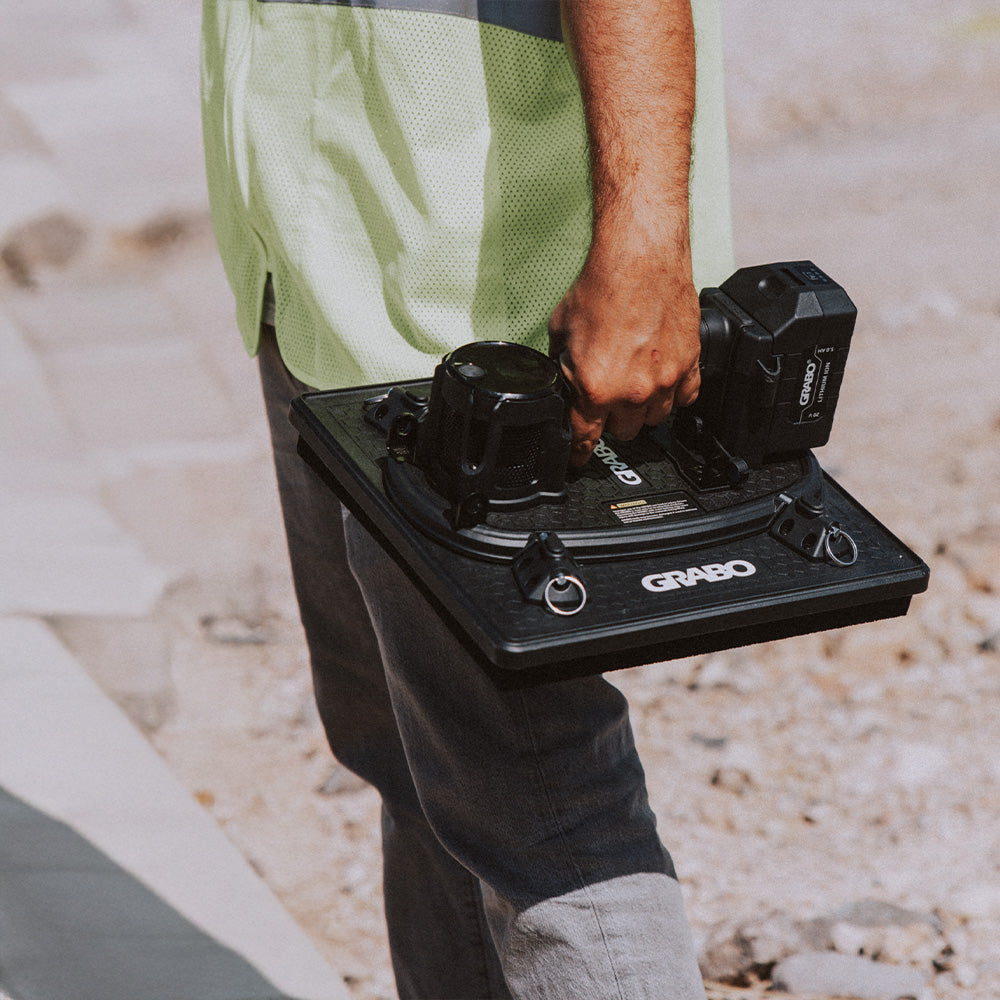 A person outdoors holding a black GRABO industrial robotic arm controller with two black end-effectors, wearing a light green high-visibility shirt and dark pants.