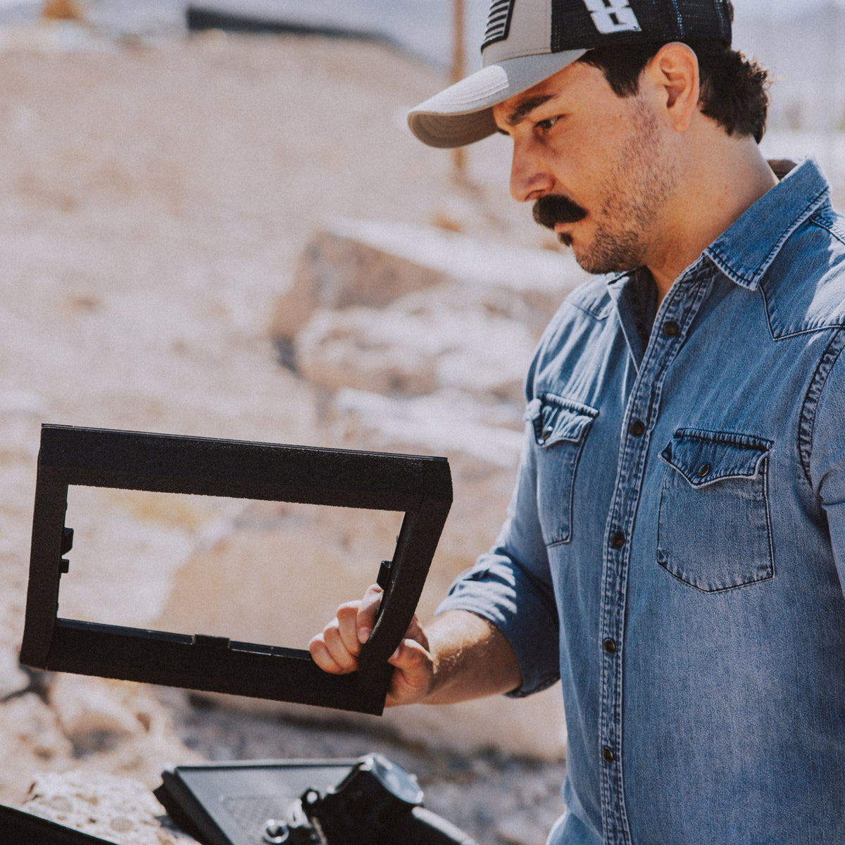 A man in a denim shirt and baseball cap holds a black rectangular frame or mounting piece outdoors in a rocky, sandy environment.