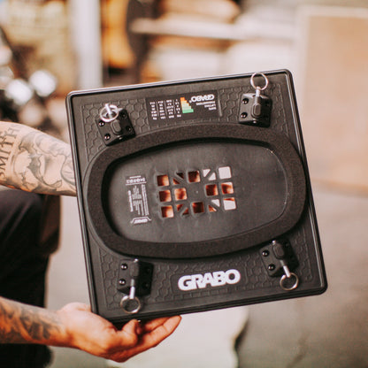 A close-up of a person holding a black GRABO  pad. The product is shown from the underside, displaying its textured black surface, mounting hardware with keys, and the white GRABO logo near the bottom.