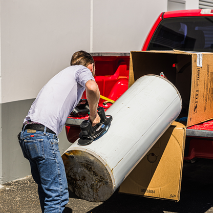 A man in a light purple t-shirt and blue jeans uses a black and blue floor jack to lift a large white cylindrical water heater out of a red pickup truck bed, with cardboard boxes nearby.