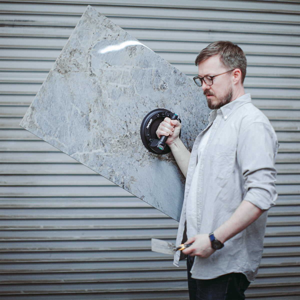 A man wearing glasses and a light gray button-up shirt holds a large, square gray marble tile with a circular mounting hole in the center, standing in front of a gray metal roll-up door.